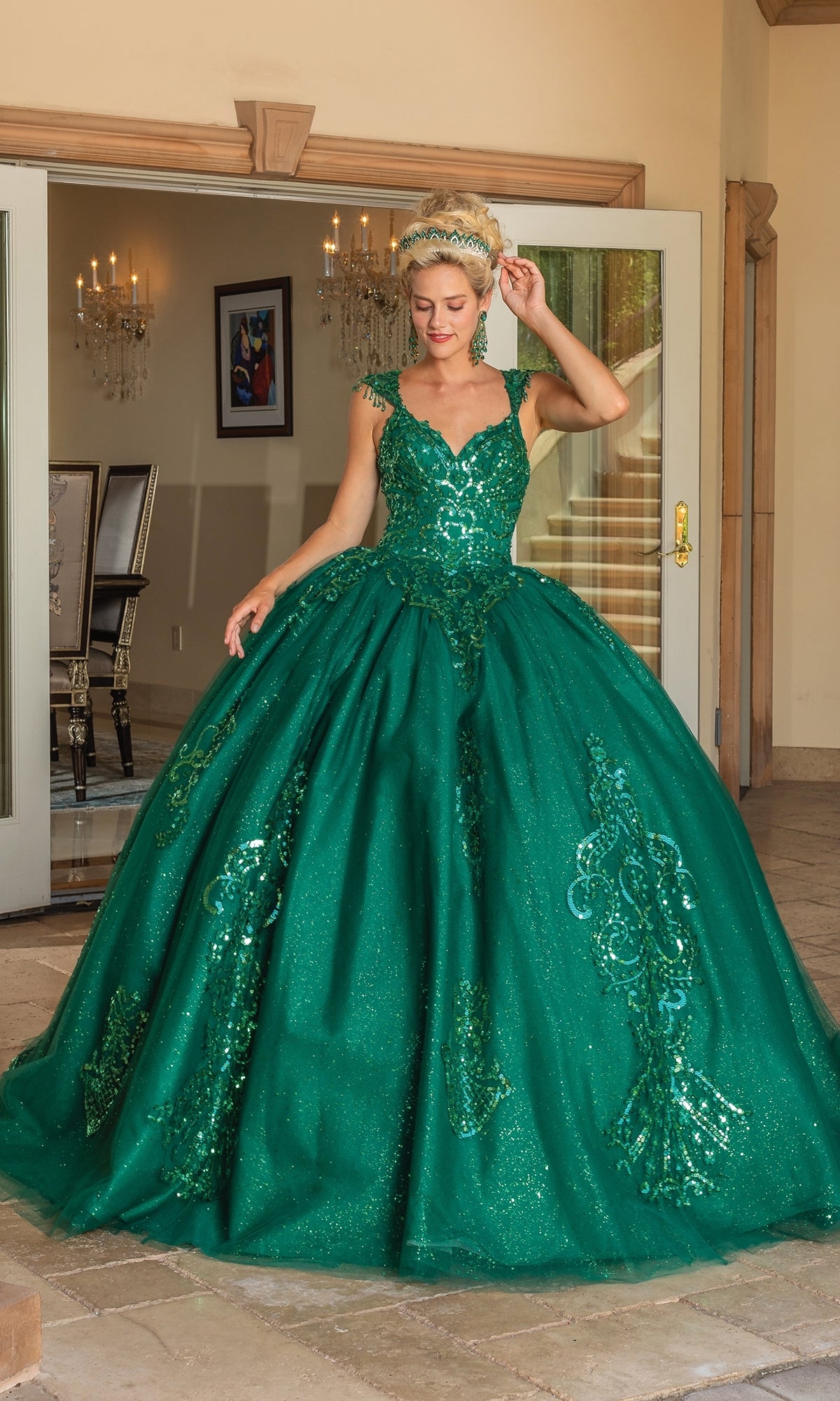 A woman poses by open glass doors, wearing the Quinceanera Dress 1758 By Dancing Queen—an emerald green A-line ball gown with sparkling glitter lace embroidery and one hand elegantly raised near her head.